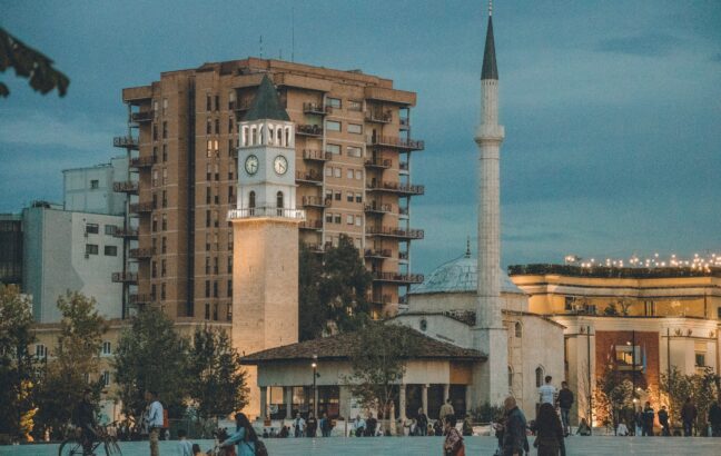 a group of people walking around a plaza with a clock tower in the background